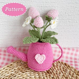 Op de afbeelding: Een handgemaakte gehaakte gieter in felroze, met een boeket gehaakte bloemen. De bloemen zijn roze en wit, met groene bladeren. Een wit hart staat op de voorkant van de gieter. Het woord "Pattern" staat in een roze ovaal.