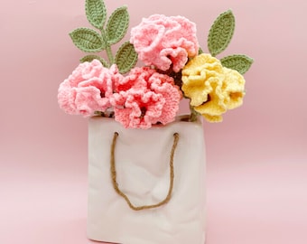 May include: A bouquet of crocheted flowers in pink, yellow, and green colors arranged in a white ceramic vase with handles.