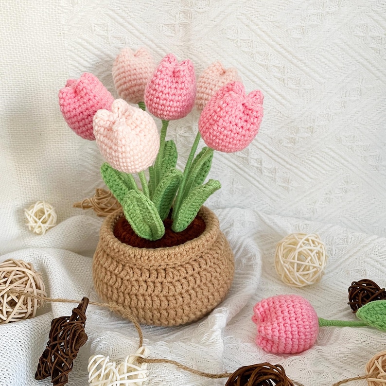 May include: A handcrafted crochet tulip arrangement in a brown pot. The tulips are in varying shades of pink, with green stems and leaves. The pot is a light brown color, and the arrangement sits on a white textured surface.