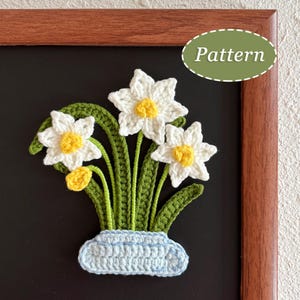 May include: Crocheted daffodil bouquet in a light blue vase, set against a black background. The flowers are white with yellow centers, and the leaves and stems are green. The word "Pattern" is in a green oval.