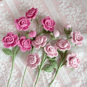May include: A bouquet of crocheted pink roses with green leaves on a white lace background.