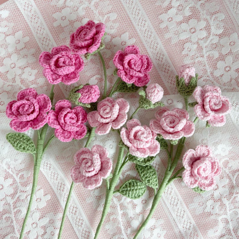 May include: A bouquet of crocheted pink roses with green leaves on a white lace background.