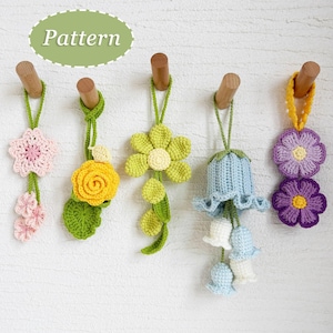 Peut inclure: Cinq ornements de fleurs au crochet faits à la main, dans des couleurs variées : rose, jaune, vert, bleu et violet. Chaque fleur est suspendue par une boucle crochetée verte ou jaune et accrochée à une cheville en bois. Le mot "Pattern" est dans un ovale vert en haut.