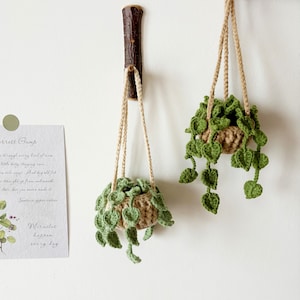 May include: Two hanging macrame plant hangers with green crocheted leaves and beige crocheted pots. The hangers are made of natural twine and are hanging from a wooden hook and a white wall.