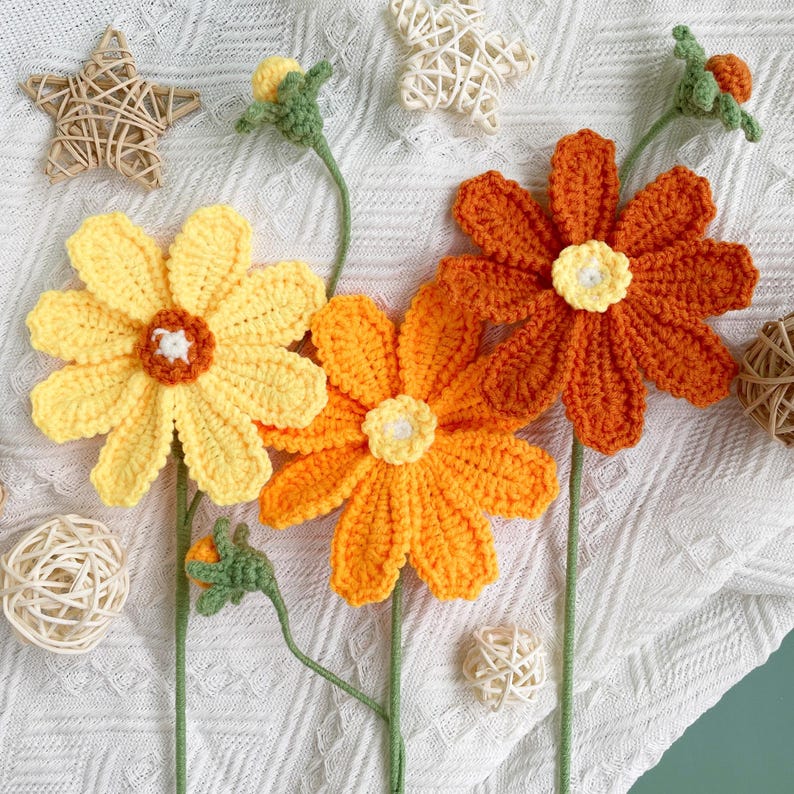 May include: Three crocheted flowers in shades of yellow and orange. The flowers are arranged on a white background with a woven star and other decorative elements.