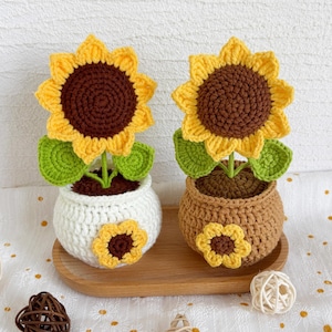 May include: Two handmade crochet sunflowers in pots. The sunflowers have yellow petals and brown centers. One pot is white with a sunflower applique, the other is brown. Green leaves and stems complete the design. The pots are on a wooden tray.