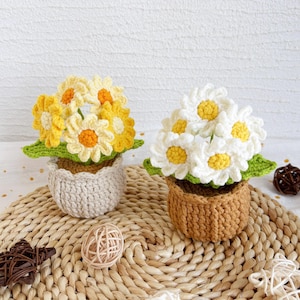 May include: Two crocheted flower arrangements in small pots. One arrangement features yellow daisies with orange centers, the other has white daisies with yellow centers. The pots are beige and brown, sitting on a woven mat.