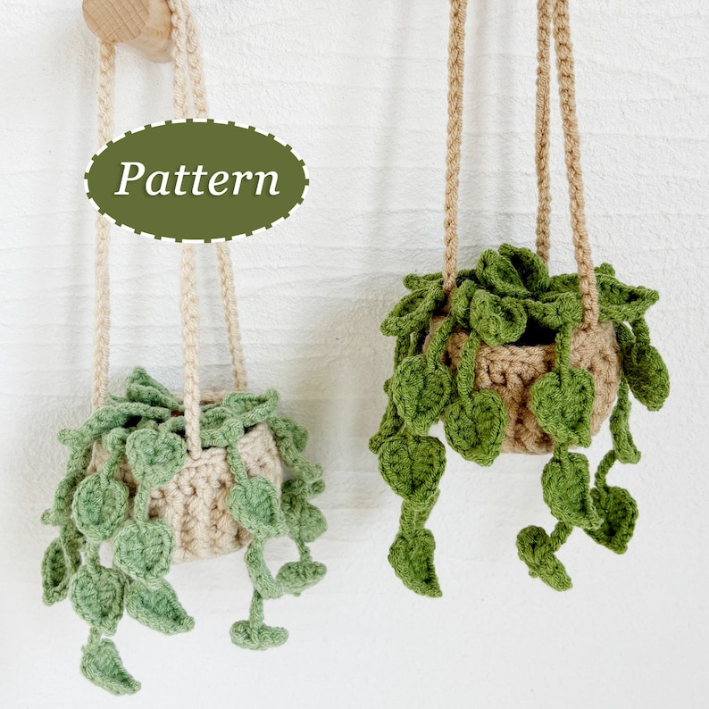 May include: Two crocheted hanging plant pots with green leaves. The pots are beige and the leaves are a bright green. The word "Pattern" is written on a green oval in the background.