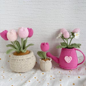 May include: Crocheted tulip flowers in pots and a watering can. The flowers are pink and white, with green leaves. The pots are beige and the watering can is pink with a heart. A charming, handmade floral arrangement.