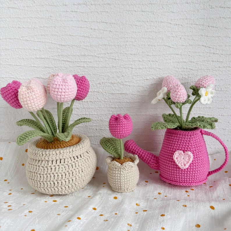 May include: Crocheted tulip flowers in pots and a watering can. The flowers are pink and white, with green leaves. The pots are beige and the watering can is pink with a heart. A charming, handmade floral arrangement.