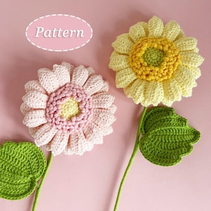 Peut inclure: Deux fleurs crochetées avec des tiges et des feuilles vertes sur fond rose. L'une est rose et jaune, l'autre jaune et vert. Le mot "Pattern" est dans un ovale rose.