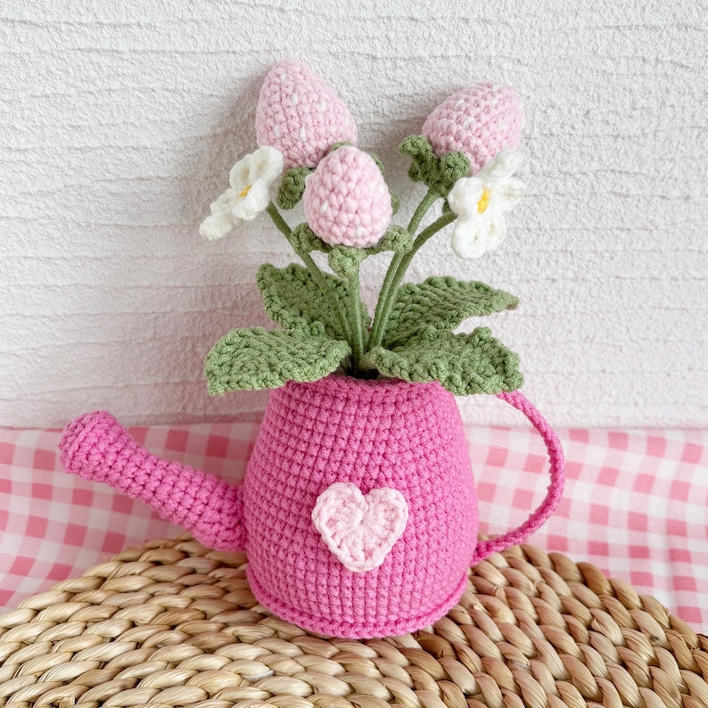 May include: A handmade crochet watering can in bright pink, adorned with a light pink heart. It holds a bouquet of crochet flowers, including pink buds, white blossoms, and green leaves. The watering can sits on a woven surface.