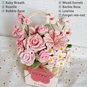 May include: A crocheted flower arrangement in a wicker basket. The flowers are pink and white and include baby's breath, roselle, bubble rose, wood sorrels, barbie rose, lewisia, and forget-me-nots. The basket has a tag that says "Wish you all the best. Be happy."