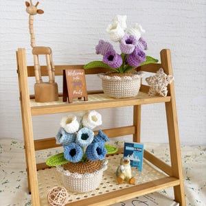 May include: A wooden shelf displays crochet flowers in pots, a wooden giraffe, a small sign that says "Welcome to the Party", and a small penguin figurine. The crochet flowers are in shades of blue, white, and purple.