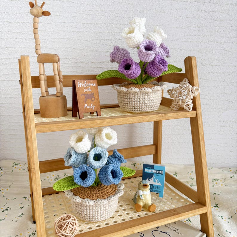 May include: A wooden shelf displays crochet flowers in pots, a wooden giraffe, a small sign that says "Welcome to the Party", and a small penguin figurine. The crochet flowers are in shades of blue, white, and purple.