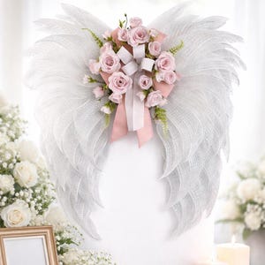 May include: A decorative angel wing wreath made of white mesh, adorned with pink roses, white ribbons, and greenery. The wings are mounted on a white surface, creating a soft, ethereal look. The wreath is a beautiful accent for memorial or wedding decor.