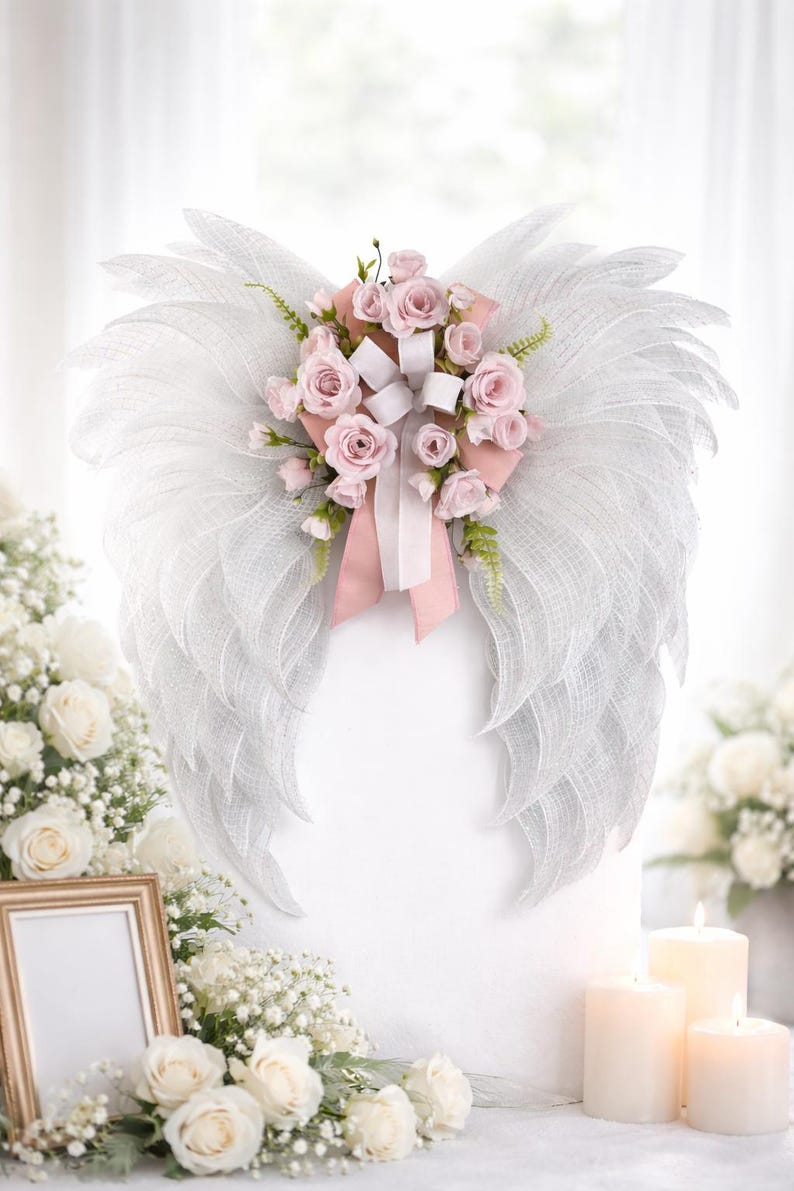 May include: A decorative angel wing wreath made of white mesh, adorned with pink roses, white ribbons, and greenery. The wings are mounted on a white surface, creating a soft, ethereal look. The wreath is a beautiful accent for memorial or wedding decor.