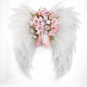 May include: White angel wings with a mesh design, adorned with pink roses, white ribbons, and greenery. The wings are a decorative item, perfect for wall decor or special events.