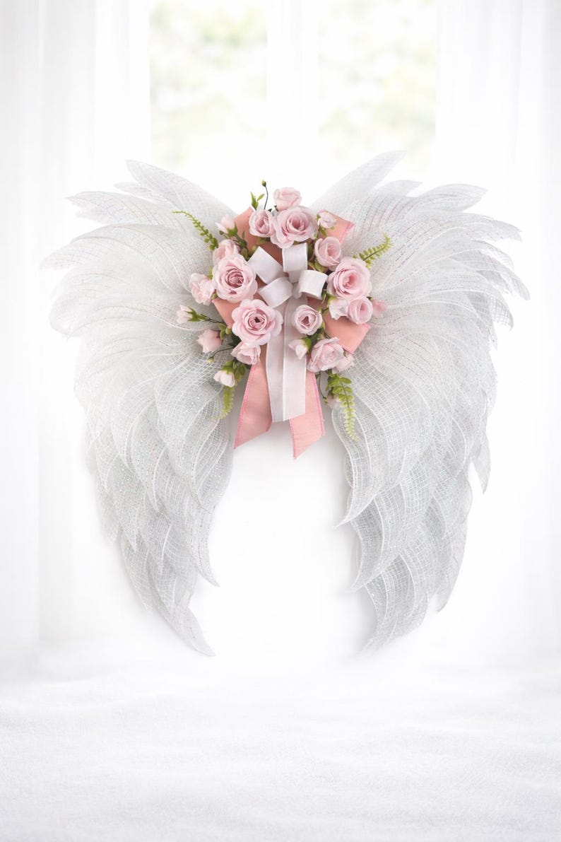 May include: White angel wings with a mesh design, adorned with pink roses, white ribbons, and greenery. The wings are a decorative item, perfect for wall decor or special events.