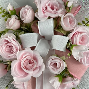 May include: A floral wreath featuring light pink roses, white ribbon, and green foliage. The roses are in various stages of bloom, from buds to fully open flowers. The wreath has a mesh backing and pink ribbon accents.
