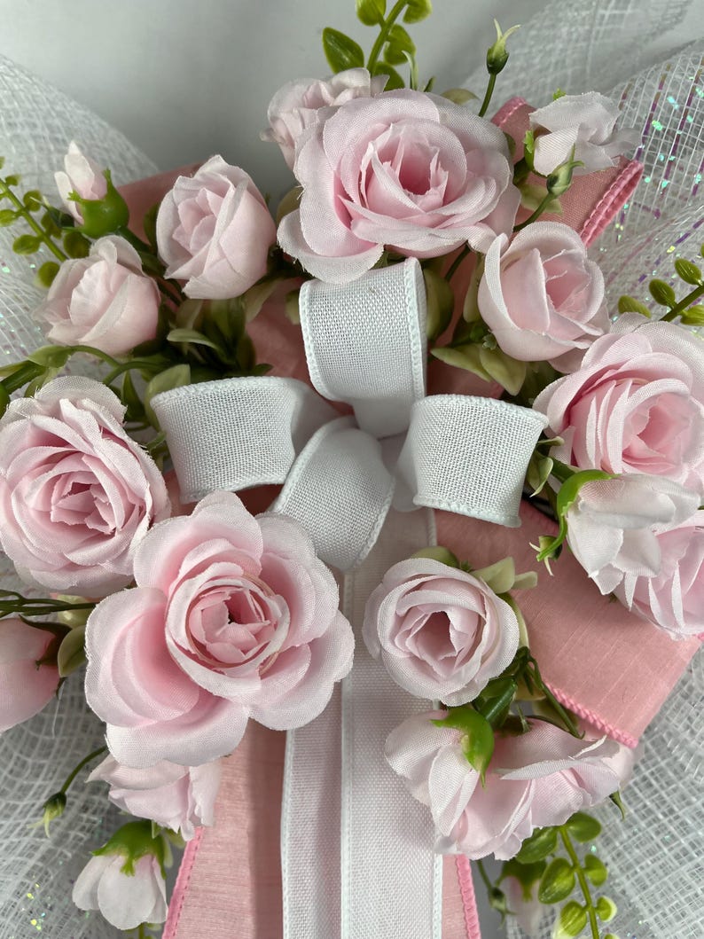 May include: A floral wreath featuring light pink roses, white ribbon, and green foliage. The roses are in various stages of bloom, from buds to fully open flowers. The wreath has a mesh backing and pink ribbon accents.