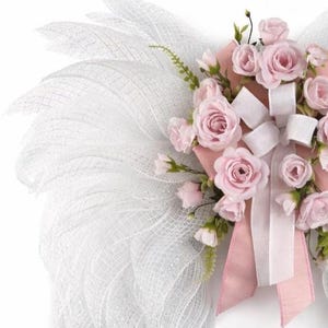 May include: A decorative wreath featuring a white mesh design and a cluster of pink roses. The roses are accented with white ribbons and a pink bow. The wreath is designed with a delicate and airy aesthetic.