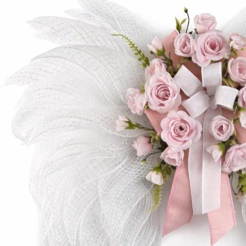May include: A decorative wreath featuring a white mesh design and a cluster of pink roses. The roses are accented with white ribbons and a pink bow. The wreath is designed with a delicate and airy aesthetic.