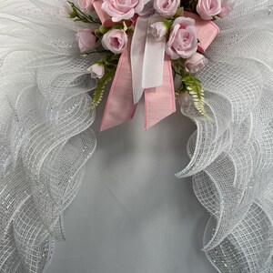 May include: A decorative angel wing wreath with white mesh wings and a cluster of pink roses, white and pink ribbons. The wreath is designed for home decor and special occasions.