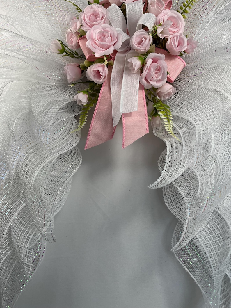 May include: A decorative angel wing wreath with white mesh wings and a cluster of pink roses, white and pink ribbons. The wreath is designed for home decor and special occasions.