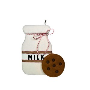May include: A white felt milk bottle with a red and white twine bow and the word "MILK" printed on the front. A brown felt chocolate chip cookie is next to the bottle.