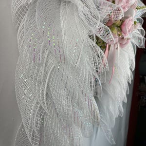 May include: A white decorative wreath with a mesh design and pink roses. The wreath features layers of white mesh with iridescent accents. Pink roses and ribbons are clustered on the right side, creating a delicate and elegant appearance.