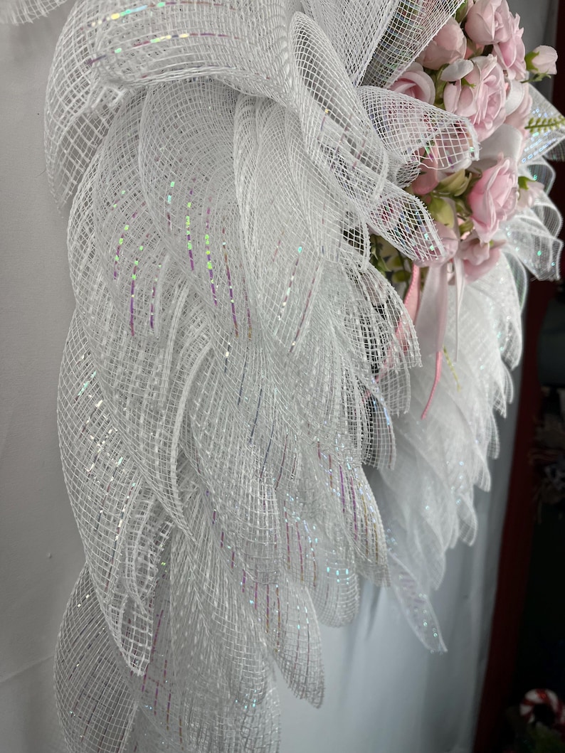May include: A white decorative wreath with a mesh design and pink roses. The wreath features layers of white mesh with iridescent accents. Pink roses and ribbons are clustered on the right side, creating a delicate and elegant appearance.