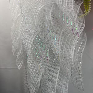 May include: A close-up of a white, layered mesh wreath with iridescent glitter accents. The wreath has a flowing, feathery appearance, with a few pink flowers and green leaves visible in the background. The mesh is tightly woven, creating a textured effect.