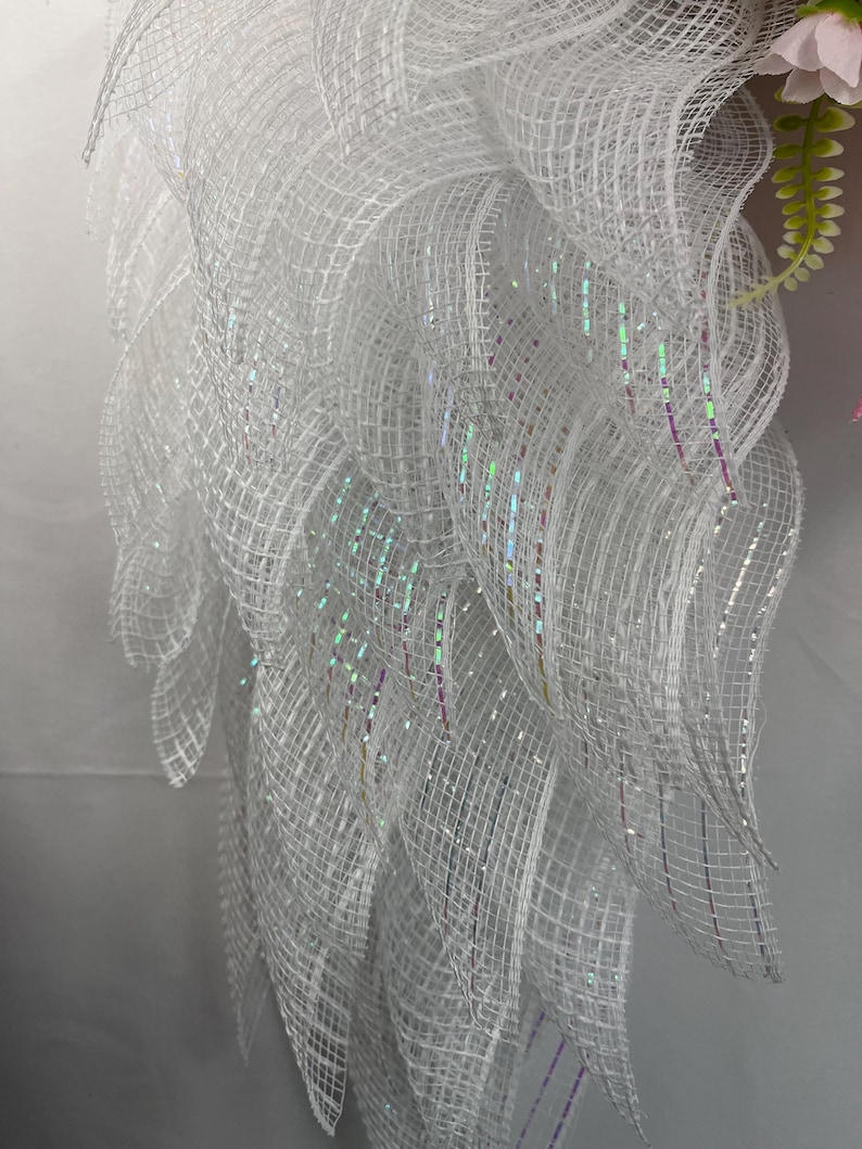 May include: A close-up of a white, layered mesh wreath with iridescent glitter accents. The wreath has a flowing, feathery appearance, with a few pink flowers and green leaves visible in the background. The mesh is tightly woven, creating a textured effect.