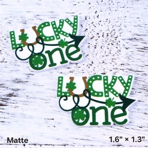 May include: Two matte white and green "Lucky One" charms with a horseshoe, shamrocks, and an arrow. Each charm measures 1.6" x 1.3". The text is in green with white accents. The background is a white, textured surface.