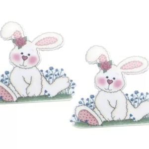 以下が含まれることがあります： ピンクの頬と頭にピンクの花をつけた白いウサギの置物2つ。緑の芝生の上に青い花が咲いています。