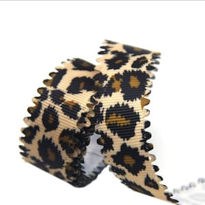 May include: A coiled length of ribbon featuring a tan background with a black and dark brown leopard print pattern. The ribbon has a distinctively textured, ribbed surface and decorative scalloped edges along both sides of the length.