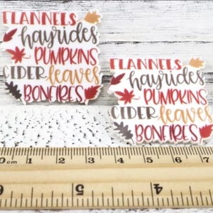 May include: Two white, fall-themed decorative stickers with the words "Flannels, hayrides, pumpkins, cider, leaves, bonfires" in various colors. A ruler with centimeter markings is at the bottom.