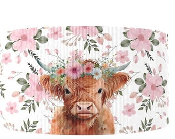 May include: A cylindrical lampshade featuring a watercolor illustration of a Highland cow wearing a floral crown. The cow is brown with long, shaggy fur. Pink flowers and green leaves are scattered across a white background.