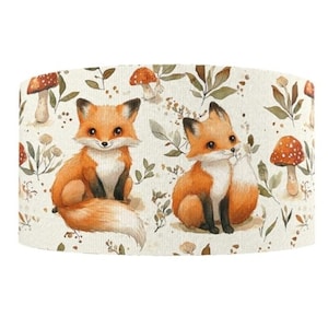 May include: A cylindrical lampshade with a watercolour design of two orange foxes with white bellies, surrounded by mushrooms and foliage. The background is cream-coloured. The foxes have black paws and ears. The mushrooms have red caps with white spots.