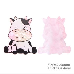Pode incluir: Duas peças decorativas em forma de vaca. Uma é uma vaca de desenho animado com corpo branco, manchas pretas e rosto rosa. A outra é uma forma de vaca rosa translúcida. O texto na imagem diz: TAMANHO: 42x50mm, Espessura: 4mm.