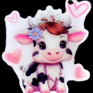 Pode incluir: Um berloque brilhante em forma de vaca com uma coroa, uma flor e corações. A vaca tem um corpo branco com manchas pretas, orelhas rosa e um nariz rosa. O berloque é rodeado por corações rosa e glitter.