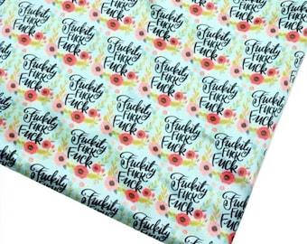May include: A light blue fabric with a repeating pattern of the word "Fuckity Fuck" in black script, surrounded by floral wreaths in red, pink, and green. The fabric is angled, showing its texture and design.