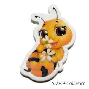 May include: A cartoon-style bee-shaped decoration with a white outline. The bee is yellow and orange with black stripes, holding a white daisy. The text "SIZE: 30x40mm" is at the bottom.