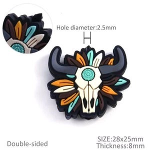 Op de afbeelding: Een dubbelzijdige schoenbedel in zwart, turquoise en bruin, met een stierschedelontwerp met hoorns en veermotieven. De bedel meet 2,8 cm x 2,5 cm en is 8 mm dik. De gatdiameter is 2,5 mm.