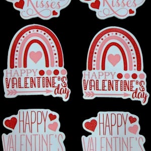 Puede incluir: Pegatinas del Día de San Valentín con diseños rojos y rosas. Las pegatinas presentan frases como "Hugs & Kisses" y "Happy Valentine's Day" con gráficos de corazones y arcoíris. Fondo negro.