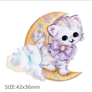 Op de afbeelding: Een wit kitten met een paarse strik zit op een gele halve maan met sterren. Het kitten heeft blauwe ogen en een roze neus. De maan is omgeven door een pluche witte wolk met regenboogaccenten. MAAT: 42x36mm