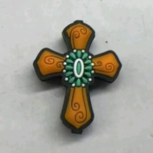 May include: A decorative orange cross charm with a green and white center. The cross has a detailed design with swirls and a beaded center.