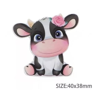 Pode incluir: Uma vaca de desenho animado com manchas pretas e brancas, usando uma flor rosa, sentada em um fundo branco. A vaca tem um nariz rosa e grandes olhos azuis. TAMANHO: 40x38mm