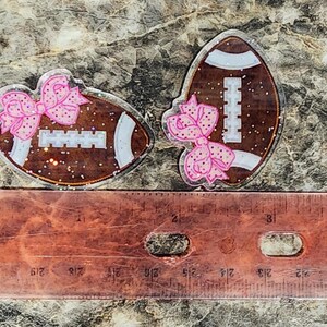 May include: Two football-shaped acrylic charms with glitter. Each charm has a brown football design with white stripes and a pink bow. The charms are next to a ruler, showing their size.